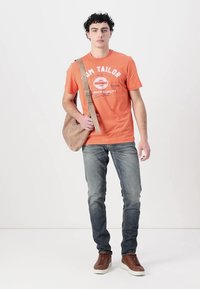 Oranža īsām piedurknēm T-kreklis ar baltu logo, kombinēts ar ziliem džinsiem un brūnām sporta apaviem. Beigā sūtīšanas soma ir uzsieta pāri plecam.