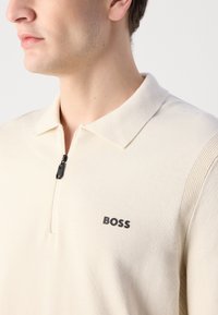 Beige polo shirt with a zippered collar, knitted texture, and embroidered "BOSS" logo in black on the chest; features ribbed accents on the shoulders.