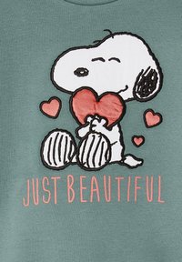 Grüne Sweatshirt mit einem gestickten Snoopy, der ein rotes Herz hält, umgeben von kleineren rosa Herzen und dem Text "EINFACH WUNDERSCHÖN" darunter.