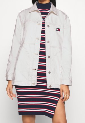 Woman wearing a light beige buttoned jacket over a navy, red, and white striped dress with a thigh-high slit on the right side.