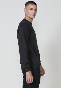 Sudadera negra texturizada con cuello redondo y diseño de panel lateral, con texto gráfico en el lado izquierdo, combinada con pantalones negros.