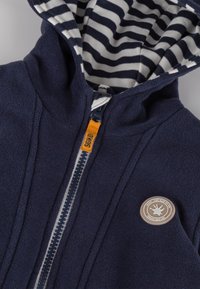 Marineblaues Fleece-Zip-Hoodie mit gestreifter Baumwollmischung innen. Enthält einen orangefarbenen Reißverschlussanhänger, ein Logo-Patch und detaillierte Nähte.