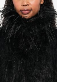 Colar de pelúcia sintética preto com fibras longas e texturizadas, criando uma aparência volumosa, complementado por uma camada inferior suave e escura.