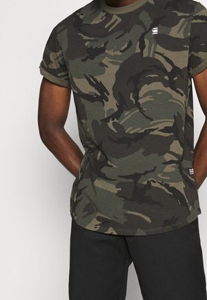 Mand iført kortærmet grøn og sort camouflage t-shirt med lille hvidt logo på venstre bryst og sorte bukser.