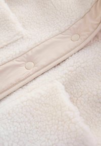Gros plan sur un tissu sherpa crème avec une bordure lisse et des boutons pression ronds sur un vêtement.
