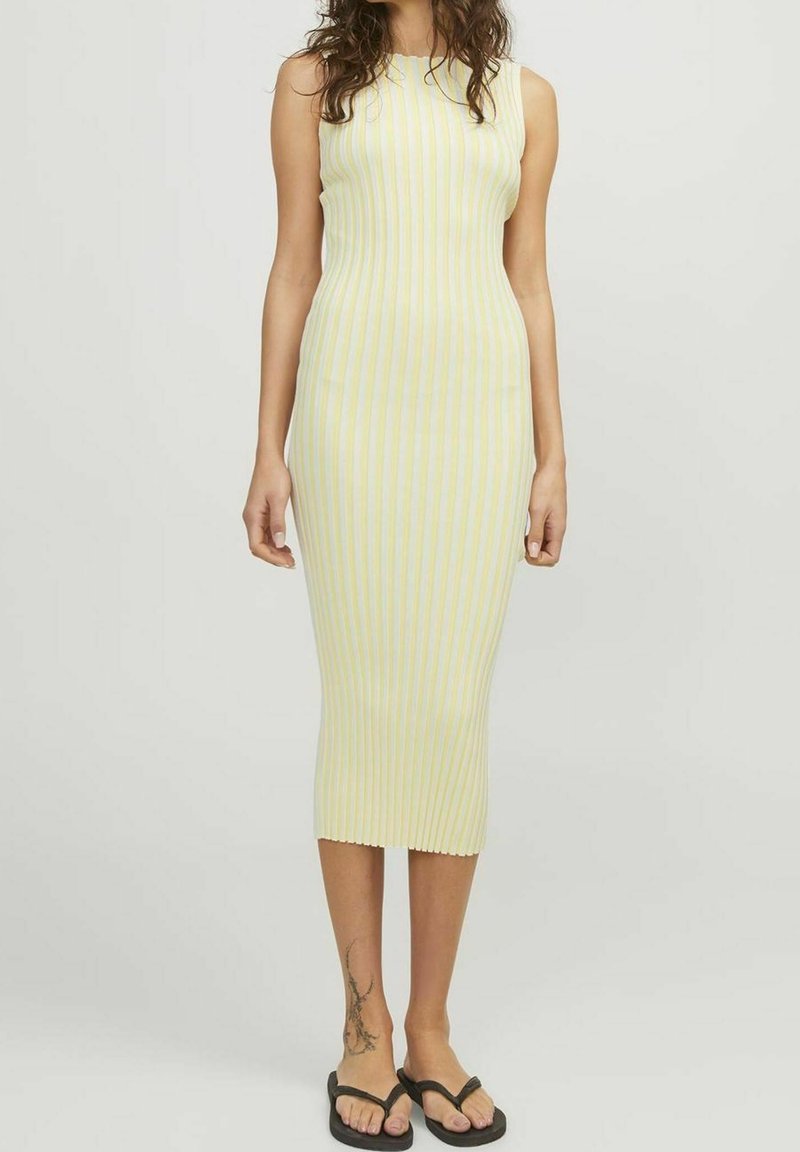Femme portant une robe midi sans manches et ajustée avec des rayures verticales jaunes et blanches, ainsi que des tongs noires, debout devant un fond uni.