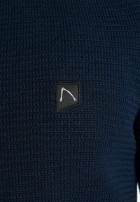 Dunkelblauer strukturierter Pullover mit einem gewebten Muster, ausgestattet mit einem kleinen schwarzen Patch, der ein weißes Design im Brustbereich zeigt.