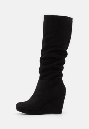 Bota de cuña de ante negra hasta la rodilla con caña caída y punta redondeada, que presenta una suela de plataforma sólida sobre un fondo blanco.