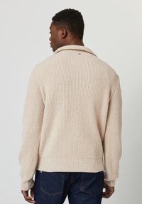 Pull en tricot côtelé beige avec un col châle et un détail de bouton à l'arrière, présentant des manches longues et une coupe ample.