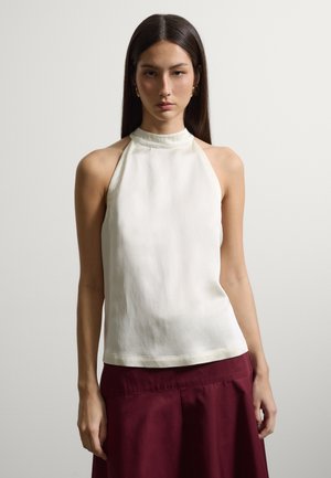 Frau mit langen dunklen Haaren trägt ein ärmelloses weißes Neckholder-Oberteil und einen burgunderfarbenen Rock und steht vor einem schlichten hellen Hintergrund.