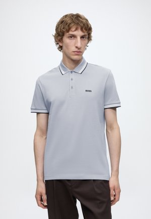 Jeune homme aux cheveux bouclés portant un polo BOSS gris clair avec une bordure noire et un pantalon foncé, debout devant un fond blanc.