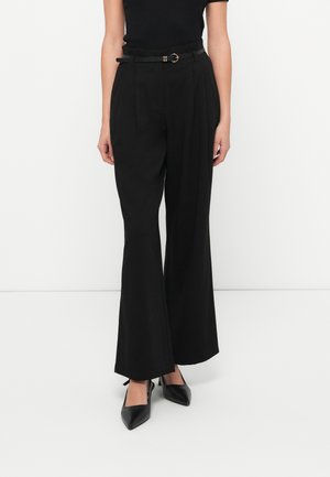 Femme portant un pantalon large noir taille haute avec ceinture et des talons noirs à bout pointu, debout devant un fond blanc uni.