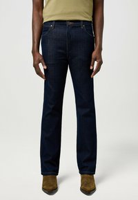 Jean en denim bleu foncé avec une coupe droite, cinq poches, coutures renforcées et fermeture à bouton. Porté avec des bottes en daim marron.