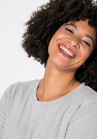 Chemise grise à manches courtes avec un col rond, texture douce et coupe décontractée ; la mannequin affiche des cheveux bouclés et un maquillage naturel, souriant.