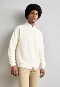 Creme pullover sweatshirt med høj hals, ribbede manchetter og et subtilt logo i lysebrune nuancer. Bæres over en hvid skjorte med krave og beige bukser.
