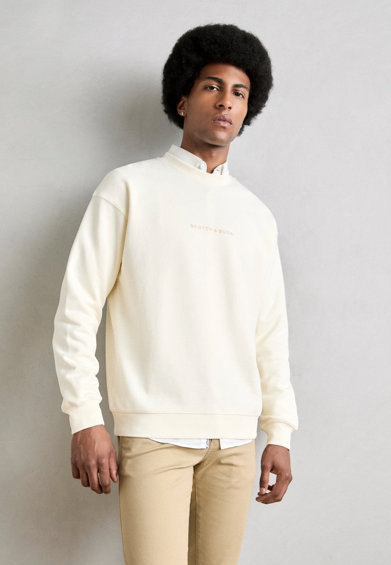 Creme pullover sweatshirt med høj hals, ribbede manchetter og et subtilt logo i lysebrune nuancer. Bæres over en hvid skjorte med krave og beige bukser.