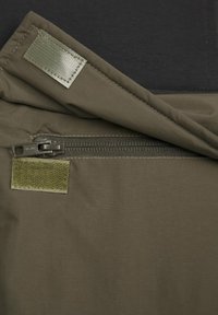 Olivgrön tyg med en slät textur, med en dragkedjeficka och ett kardborrefäste. Bakgrunden är svart, vilket visar en kontrasterande design.