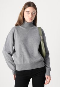 Pull en col roulé gris en matériau doux. Présente des poignets et un ourlet côtelés. Assorti à un sac à épaule vert, sur fond blanc uni.