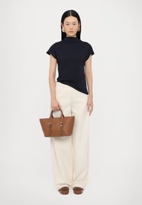Top a maniche corte blu navy, pantaloni larghi color crema e borsa in pelle marrone con hardware dorato, in piedi su uno sfondo uniforme.