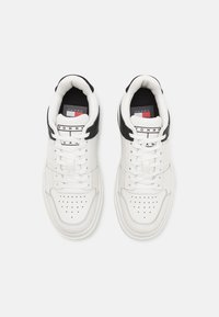 White and black lace-up sneakers with perforated toe caps and Tommy Jeans branding on tongue and insole, shown from above on white background.