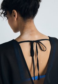 Black sheer garment with a deep V-back, featuring a tied collar accent and a blue strap visible underneath. Textured fabric with subtle sheen.