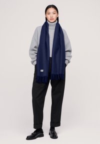 Écharpe en laine navy avec des bords frangés, drapée sur un pull à col roulé gris. La tenue est complétée par un pantalon noir ajusté et des chaussures.