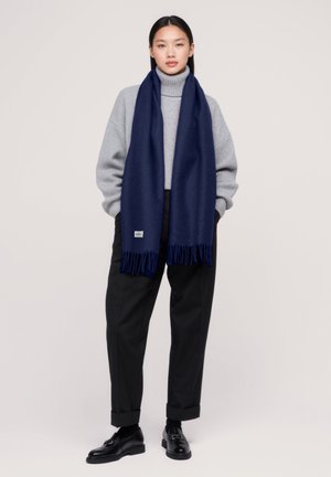Sciarpa di lana blu navy con bordi frangiati, appoggiata su un maglione grigio a collo alto. L'outfit comprende pantaloni neri a sigaretta e scarpe.