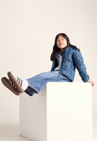 Young girl in blue quilted jacket and jeans sitting on a white cube, legs extended, against a plain light background.