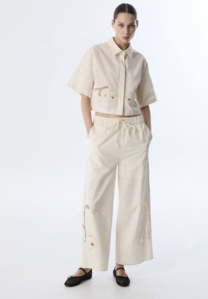 Camisa corta de color crema con mangas cortas y detalles recortados, combinada con pantalones de pierna ancha que presentan acentos recortados similares.