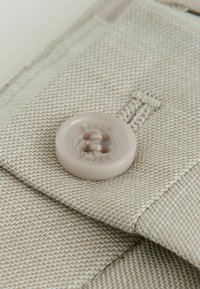 Svetlo bež tkanina s ribjo kostjo, z velikim, gladkim, svetlo roza gumbom s štirimi luknjami in detajlom šiva.