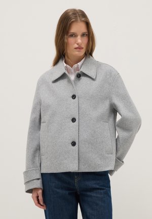 Vrouw draagt een lichtgrijze jas met knopen, een brede kraag en manchetdetails over een gestreept overhemd en donkerblauwe spijkerbroek.