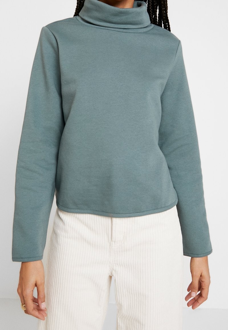Personne portant un pull col roulé vert discret et un pantalon en velours côtelé blanc cassé, debout les bras détendus le long du corps.