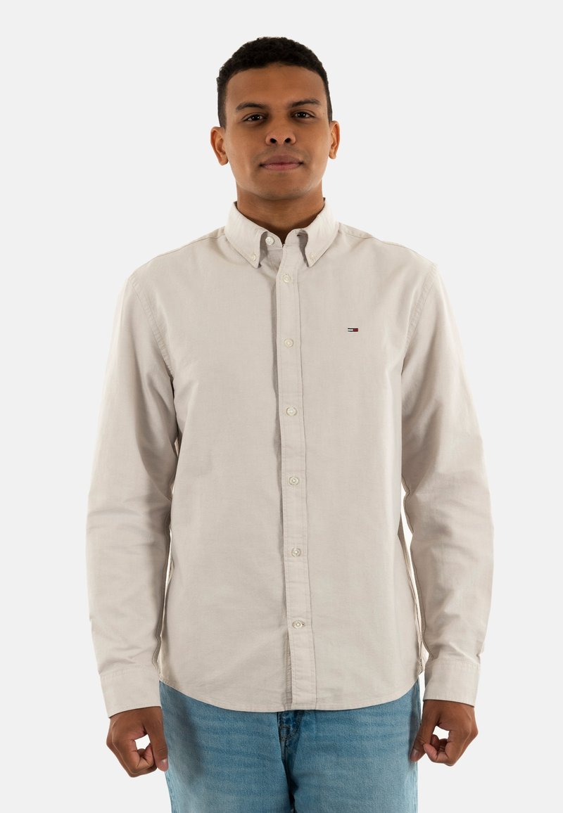 Camicia a maniche lunghe color beige chiaro con colletto, chiusura con bottoni e dettagli del logo discreti sul lato sinistro del petto, abbinata a pantaloni jeans blu chiaro.