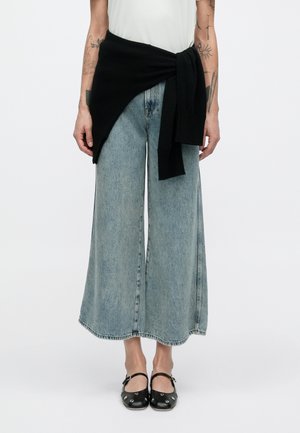 Široké nohavice z ľahkého modrého denimu s vyblednutým efektom, skombinované s čiernym pleteným svetrom zviazaným v páse a čiernymi nazúvacími topánkami s detailmi.