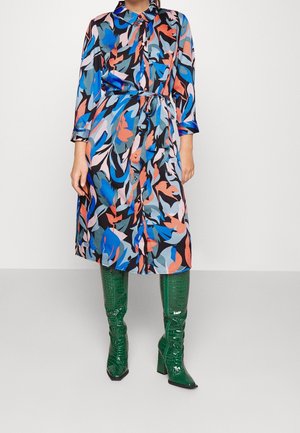 Kvinde iført en farverig kjole med abstrakt mønster og bindebånd i taljen samt grønne knælange teksturerede støvler, stående foran en ensfarvet baggrund.