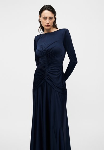 Γυναίκα με σκούρα μαλλιά σε μακρυμάνικο φόρεμα σε ναυτικό μπλε με ραφές στο μπούστο, στέκεται μπροστά σε ένα απλό φωτεινό φόντο.