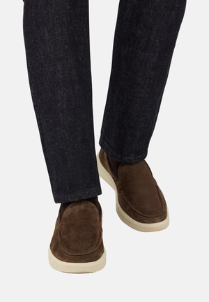 Zapatos marrones de ante sin cordones con punta redonda y suela beige, combinados con jeans de mezclilla oscuros, mostrando una silueta casual y un contraste de texturas.