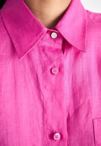 Ljusa rosa linne skjorta med krage, med en knäppning fram, två knappar på knäppningen, och en bröstficka. Slät textur.