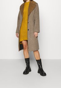 Cappotto lungo a quadri marrone con colletto in camoscio marrone, abbinato a un vestito lavorato a maglia giallo senape e stivali neri con tacco spesso.