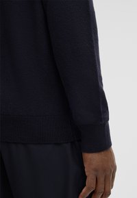 Nahaufnahme einer Hand und des Unterarms einer Person, die einen dunkelblauen Strickpullover und passende dunkle Hosen tragen, vor einem schlichten hellen Hintergrund.