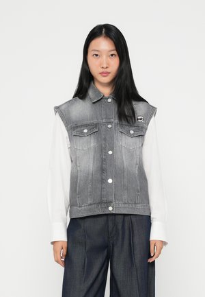 Szürke denim ujjatlan dzseki elülső zsebekkel, fémgombokkal és enyhe ombre hatással. Fehér hosszú ujjú póló felett viselve.