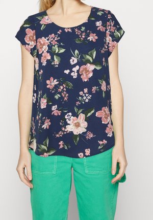 Femme portant un haut bleu marine à manches courtes avec imprimé floral rose et un pantalon vert vif avec de grandes poches avant.