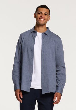 Lichtblauw overhemd van zachte stof, met lange mouwen en een standaardkraag, gelaagd over een wit t-shirt, gedragen met donkere broeken.
