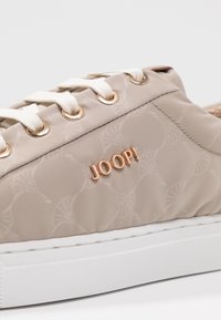 Baskets beige décontractées avec lacets blancs, œillets dorés, motif floral subtil et logo métallique en relief "JOOP!" sur le côté.