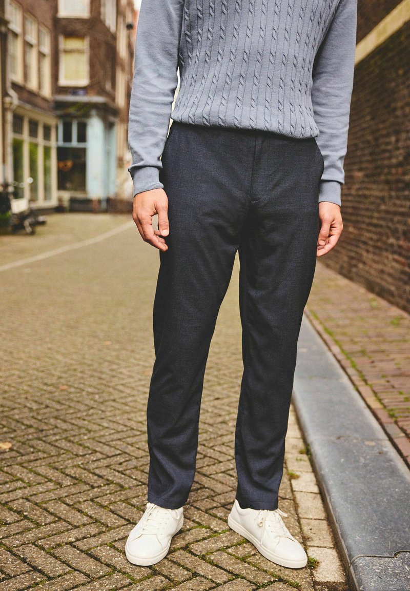 Lichtgrijze kabelgebreide sweater, donkere tapered broeken en witte sneakers. De outfit heeft een gestructureerd patroon en een getailleerd ontwerp.