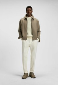 Olivgrön jacka, krämfärgad texturerad tröja, vita skräddarsydda byxor och beige sneakers. Slät tyg med strukturerad design och subtil färgkontrast.