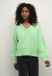 Lichtgroene, oversized trui van zachte, gestructureerde garen; heeft een v-hals, geribbelde zoom en ballonmouwen. Gepaard met donkere jeans.