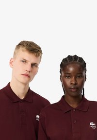 Bordowe koszulki polo o teksturowanym wykończeniu, z trzema guzikami, kołnierzykiem oraz białym haftowanym logo Lacoste na lewej piersi.