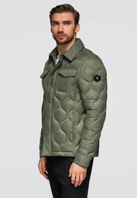 Uomo che indossa una giacca trapuntata verde oliva con tasche sul petto a patta e pantaloni neri, in piedi di lato contro uno sfondo neutro.