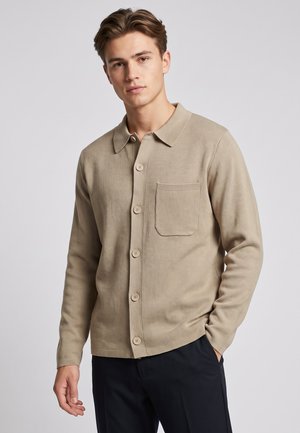 Junger Mann, der eine beige Knopfleiste Strickjacke mit Brusttasche und schwarze Hose trägt, vor einem schlichten hellen Hintergrund stehend.